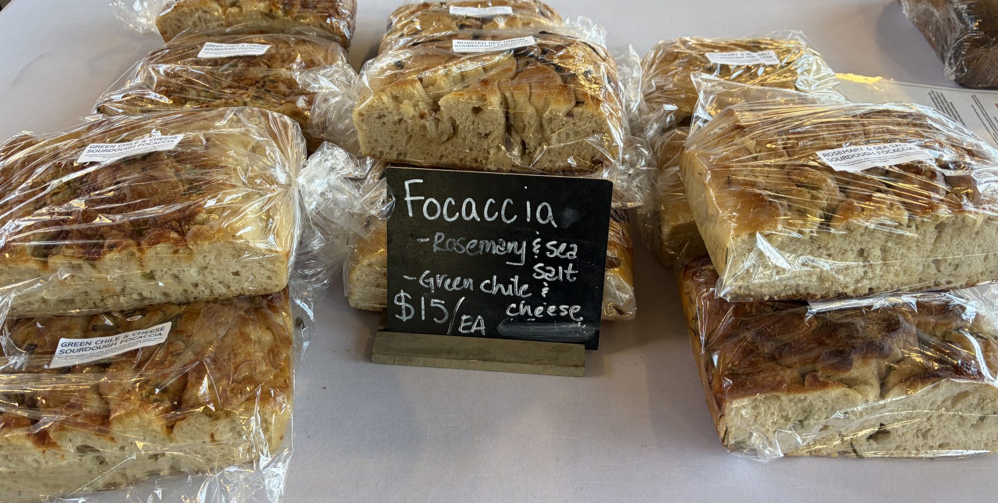 A market table of bagged sourdough focaccia loaves behind a chalkboard reading Focaccia, rosemary and sea salt, green chile and cheese, $15 each