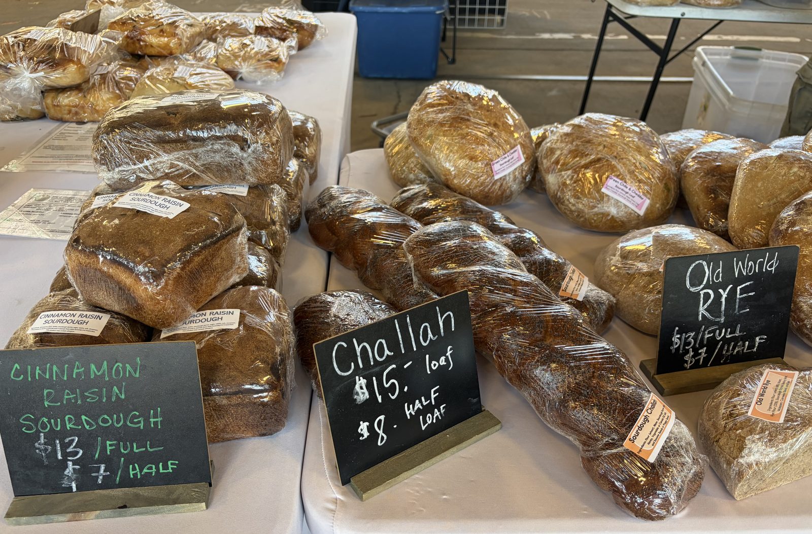Market table of bagged sourdough loaves with chalkboards reading Cinnamon Raisin Sourdough $13 full $7 half, Challah $15 loaf $8 half loaf, and Old World Rye $13 full $7 half