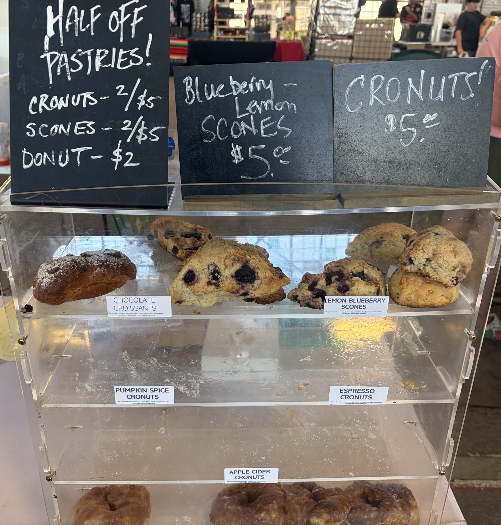 Clear pastry case with chalkboards reading Blueberry-Lemon Scones $5 and Cronuts $5, and a Half Off Pastries board; case labels read chocolate croissants, lemon blueberry scones, pumpkin spice cronuts, espresso cronuts and apple cider cronuts