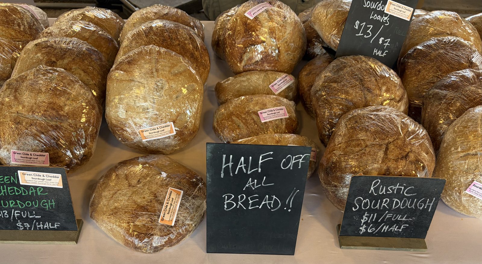 Rows of bagged Green Chile & Cheddar sourdough loaves with chalkboards reading $13 full $7 half, Rustic Sourdough $11 full $6 half, and a board reading Half Off All Bread