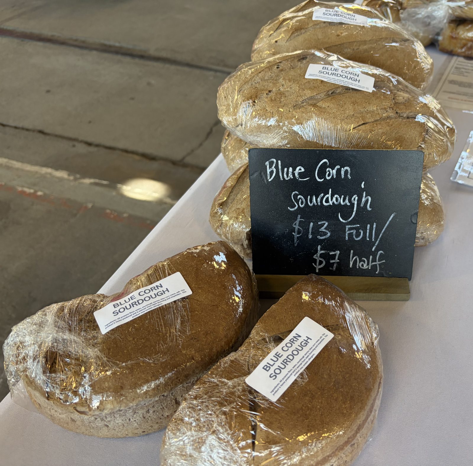 Bagged blue corn sourdough loaves beside a chalkboard reading Blue Corn Sourdough $13 full $7 half; bag labels list all purpose flour, water, sourdough starter, blue corn masa and salt